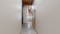 This character-rich urban loft showcases a thoughtful conversion of a historic building, blending industrial heritage with modern livability across a single floor. The open-concept living, dining, and kitchen area serves as the heart of the space, where exposed red brick walls, original heavy timber beam ceilings, and polished concrete floors create a striking industrial aesthetic. Large windows along the brick wall flood the living area with abundant natural light, while a ceiling fan adds practical comfort. The kitchen is outfitted with light maple cabinetry, dark granite countertops, stainless steel appliances including a range and over-the-range microwave, and a peninsula that defines the cooking zone. Hallways connect the living area to two bedrooms, each featuring the same concrete flooring and wood beam ceilings, with one room incorporating a built-in platform element. The two bathrooms include a full bath with a distinctive curved walk-in shower flanked by glass block panels, and a second bath featuring a double-sink maple vanity with a dark countertop and matte black fixtures. Additional utility and laundry rooms, along with two closets, provide practical storage. Exposed structural columns and industrial conduit throughout reinforce the loft's authentic, converted character.