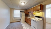 This single-floor unit is designed with practicality in mind, featuring consistent drop-panel ceilings and neutral wall tones throughout. The kitchen serves as a central functional space, equipped with warm oak cabinetry offering ample upper and lower storage, a tile backsplash in muted yellow and beige tones, and white appliances including a coil-burner electric range, refrigerator, and a single-basin sink. Tile flooring runs through the kitchen, providing a durable, easy-to-clean surface, while a wall-mounted air conditioning unit and baseboard heating ensure year-round climate control. Natural light enters through a window fitted with horizontal blinds.

The remaining rooms feature patterned carpeting in a neutral beige tone, creating a softer underfoot feel. Each room benefits from natural light via windows with blinds and is equipped with wall-mounted heating units. The spaces are clean and open, providing flexibility for various uses.

The bathroom includes full-surround square tile walls in a warm beige, a soaking bathtub with a shower, a toilet, and a vanity with counter space. A window allows natural light into the room. The overall ambiance of the unit is understated and functional, suited for straightforward, comfortable living.