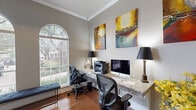 Located at 2742 Jeanetta Rd in Houston, TX, this thoughtfully designed office space offers a refined and functional environment suited for focused, professional work. The room is anchored by a continuous granite-topped built-in desk that wraps along the walls, providing generous workspace for two workstations equipped with ergonomic mesh chairs. A set of elegant white built-in shelves with glass-fronted lower cabinets offers ample storage and display space, adding both practicality and visual appeal.

The flooring throughout is rich hardwood, which complements the neutral gray walls and crisp white crown molding and baseboards. A standout architectural feature is the trio of tall arched windows along one wall, adorned with white plantation shutters that allow for controlled natural light and outdoor views of the surrounding greenery. A French door with transom window provides a sophisticated entry point, further enhancing the room's classic character.

Decorative touches, including colorful abstract canvas paintings and ornamental metal wall art, lend a creative and vibrant energy to the workspace. Task lamps with black shades provide focused lighting at each workstation. The overall ambiance strikes a balance between professional functionality and polished aesthetic, making this office a highly practical and visually engaging space.