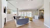Located at 1889 W Queen Creek Rd in Chandler, Arizona, this single-story community clubhouse offers a thoughtfully designed gathering space for residents. The open-concept main lounge features warm wood-look flooring, soaring ceilings with recessed lighting, and an abundance of natural light streaming through large windows and glass doors that frame views of the outdoor pool area. A tiled fireplace with a wall-mounted TV serves as a focal point, surrounded by curved sectional sofas and vibrant yellow accent chairs that lend a contemporary, welcoming feel. A built-in workstation along one wall provides a convenient workspace within the lounge. The leasing office area is bright and professional, equipped with plantation shutters, wood-tone desks, and colorful abstract artwork. A cozy lounge nook, visible from a glass partition, connects seamlessly to the fitness center, which features spin bikes and strength training equipment. The clubhouse kitchen is outfitted with stainless steel appliances, gray shaker cabinets, a herringbone tile backsplash, and a white countertop island — ideal for community events. Throughout the space, a cohesive color palette of warm grays, whites, and yellow-green accents creates a bright, modern ambiance that is both functional and inviting.