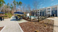 Located at 6601 Roxboro St in Raleigh, North Carolina, this outdoor amenity space is thoughtfully designed to provide a resort-style living experience. At the heart of the area is a generously sized swimming pool with sparkling blue water, framed by a spacious concrete sun deck lined with lounge chairs and navy blue umbrellas — ideal for relaxing under the North Carolina sun. A dedicated pool house structure sits at the edge of the deck, providing convenient access to poolside amenities. The pool area is enclosed by sleek black iron fencing, with landscaped mulch beds adding a polished, well-maintained aesthetic throughout.

Adjacent to the pool, a lush green lawn area features freestanding hammocks, offering a tranquil retreat backdropped by a natural woodland tree line. Additional outdoor seating and dining areas with umbrella-shaded tables provide communal gathering spaces for residents. A modern farmhouse-style clubhouse anchors one end of the property, featuring board-and-batten white siding, large glass doors, and a small outdoor activity zone with a hanging chair. Well-lit pedestrian pathways wind through the grounds, connecting the multi-story residential buildings — clad in classic grey and blue siding with private balconies — to all amenity areas, creating a cohesive and inviting community environment.