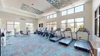 Located at 2511 Watermark Ter in Elgin, IL, this fitness center offers a thoughtfully designed, two-room exercise facility that balances functionality with modern aesthetics. The main exercise room is defined by soaring ceilings adorned with decorative teal wave-patterned ceiling panels, creating a visually striking ambiance. Large, multi-pane windows line the walls, flooding the space with abundant natural light and offering views of the surrounding community. The room features vibrant teal and gray patterned carpet tiles and houses a comprehensive array of equipment, including a row of treadmills, elliptical trainers, stationary bikes, and a full strength-training section complete with cable machines, a dumbbell rack, and weight benches. Floor-to-ceiling mirrors along one wall visually expand the space and support strength training. A vending machine provides convenient on-site refreshments. The secondary room serves as a dedicated studio space, finished with warm wood-look luxury vinyl plank flooring and illuminated by modern linear pendant lighting. This room is equipped with cycling bikes, yoga mats, and other flexible fitness accessories, along with a wall-mounted flat-screen TV — ideal for guided workouts or group classes. A glass-paneled door connects the two rooms, maintaining an open, airy feel throughout the entire facility.
