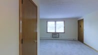 This single-floor space presents a straightforward and functional layout. The main room visible upon entry is finished with wall-to-wall carpet in a neutral tone, contributing to a quiet and understated atmosphere. A wood-paneled interior door with a brass-toned knob frames the entryway, adding a warm, traditional touch to the space.

Natural light enters through a double-pane window fitted with a wood-trimmed blind, positioned along the far wall. Beneath the window sits a wall-mounted air conditioning unit, providing in-room climate control. A baseboard heating unit runs along the base of the exterior wall, offering an additional source of warmth during cooler months.

The walls are finished in a soft, neutral tone, and the ceiling features a subtle textured finish. The room is open and unobstructed, allowing for flexible furniture arrangements. Overall, the space offers a clean, modest environment with practical built-in climate control features suited for year-round comfort.