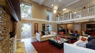 This impressive residence is defined by its dramatic two-story vaulted ceilings in the main living area, creating an expansive, airy atmosphere that immediately captures attention. Tall, floor-to-ceiling windows line the walls of the living room, flooding the space with generous natural light and offering pleasant views of the lush, tree-lined surroundings outside. The living area is thoughtfully appointed, featuring a warm, neutral color palette with tan walls accented by crisp white trim and structural columns that add architectural character. A decorative pendant light fixture anchors the room, adding an elegant focal point beneath the soaring ceiling. The interior balcony overlooking the main living space adds a distinctive architectural layer, complete with a classic wooden railing that enhances the home's open, connected feel. Beyond the living room, a dedicated workspace or study area is visible, outfitted with a desk setup framed by additional columns and warm wood tones. The flooring transitions from large-format stone tile near the entry to richly colored area rugs in the seating area, grounding the space with texture and warmth. Overall, the home presents a harmonious blend of openness, natural materials, and thoughtful design details that create an inviting and functional living environment.