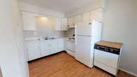 This single-floor unit offers a practical and straightforward layout across one bedroom and one bathroom. The entry opens to a spacious living area finished with wall-to-wall carpet, neutral-toned walls, and multiple windows that allow natural light to fill the space. Baseboard heating runs along the perimeter, and a wall-mounted air conditioning unit provides additional climate control. A dedicated entryway with hardwood-style flooring transitions smoothly into the living space.

The kitchen is situated off the main living area and features white cabinetry with ample upper and lower storage, light countertops, and hardwood-style flooring. White appliances include a refrigerator, electric range, and dishwasher, all arranged in an efficient L-shaped configuration.

The bedroom is a well-proportioned, carpeted room with neutral walls and a closet equipped with built-in shelving, offering functional storage.

The bathroom includes a full tub-shower combination with a classic pink-and-white tile surround, mosaic tile flooring, a vanity with sink, and white wall tile accents, giving it a clean, retro aesthetic throughout.