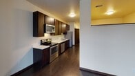 This single-floor unit offers a clean, modern design with consistent dark hardwood-style flooring and warm neutral walls throughout. The open kitchen is equipped with a full suite of stainless steel appliances—including a range, microwave, dishwasher, and refrigerator—complemented by rich dark cabinetry, light countertops, and ample upper and lower storage. The main living area is bright and spacious, featuring a large sliding glass door that opens directly onto a private balcony, allowing for generous natural light and outdoor views of the surrounding neighborhood. A secondary window further enhances the airy atmosphere of the room. The bathroom includes a wide vanity with a white countertop, an undermount sink, dark cabinetry below, and a large wall mirror with overhead lighting, creating a clean and functional space. One of the bedrooms features carpeted flooring and direct access to a walk-in closet with built-in shelving, along with a wall-mounted electrical panel. Dark door trim and baseboards provide a cohesive design accent throughout the unit. The overall layout, visible in the dollhouse view, is efficient and well-organized, offering a comfortable and functional living environment.