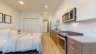 This thoughtfully designed studio unit offers a well-organized open-plan layout that efficiently integrates living, sleeping, dining, and cooking functions within a single, cohesive space. Light hardwood flooring runs throughout, lending warmth and visual continuity across the entire unit.

The kitchen is positioned along one wall and features stainless steel appliances, including a gas range, over-the-range microwave, refrigerator, and dishwasher. Two-tone cabinetry pairs white upper cabinets with warm wood-finish lower cabinets, while a white herringbone tile backsplash and light gray countertops add refined detail. Recessed lighting provides consistent illumination throughout.

The sleeping area accommodates a full-sized bed alongside a built-in closet with paneled doors, offering practical storage. Adjacent to the sleeping area, a dedicated sitting nook is anchored by expansive floor-to-ceiling windows that frame striking urban skyline views, flooding the space with natural light.

The bathroom is clean and modern, featuring a wood-tone vanity with a white countertop, undermount sink, large mirror, and a glass-enclosed shower, all finished in a crisp, neutral palette that complements the overall aesthetic of the unit.