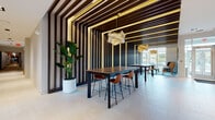 Located at 67 Old Amboy Rd in Old Bridge, New Jersey, this single-floor amenity space delivers a polished, contemporary experience through carefully curated design elements and versatile social areas.

The entrance sets a strong first impression with large-format light tile flooring and a dramatic, large-scale mosaic wall sculpture crafted from textured metallic tiles, establishing an artistic tone from the moment of arrival.

Moving deeper into the space, an expansive open lounge area features soaring double-height ceilings lined with floor-to-ceiling glass walls and oversized doors that frame outdoor views and flood the interior with natural light. Light hardwood flooring anchors the space, complementing a comfortable sectional seating arrangement and high-top café tables.

A dedicated game room offers a full-size billiards table set against a richly textured copper-toned accent wall, while a separate recreation area features a shuffleboard table alongside lush potted plants and direct access to an outdoor patio.

A standout co-working or dining corridor showcases a dramatic design treatment: dark vertical wood slat walls and ceilings integrated with warm gold linear lighting create a bold, tunnel-like visual effect, completed by sculptural pendant lights and walnut high-top tables with cognac leather bar stools. A compact kitchenette nook with a navy blue surround, open shelving, and a bistro table rounds out the amenities, providing a casual refreshment station.