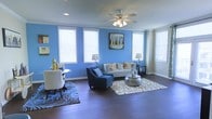 This single-floor space makes a strong visual impression through its bold blue accent wall, which serves as a defining architectural feature of the main living area. Dark hardwood flooring runs throughout, providing a rich contrast against the vivid wall color and lending warmth and cohesion to the space. Multiple windows fitted with white horizontal blinds line the walls, allowing generous natural light to fill the room while maintaining privacy. A ceiling fan with integrated lighting is centrally positioned, contributing to both air circulation and ambient illumination. The living area is generously proportioned, comfortably accommodating a sofa, accent chair, and a metallic round coffee table atop a patterned area rug. A smaller dining or accent table with seating is positioned near the windows, creating a versatile secondary zone within the same open layout. Recessed lighting complements the ceiling fan, ensuring even illumination throughout. Neutral wall tones on adjacent surfaces balance the boldness of the blue accent wall, creating a visually dynamic yet cohesive environment. Overall, the space offers an open, airy feel with a contemporary aesthetic.