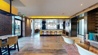 Located in Baltimore's vibrant 21231 zip code, this thoughtfully designed clubhouse spans a generous single floor, blending industrial character with modern comfort throughout its open, well-appointed layout. Wide-plank reclaimed hardwood floors anchor the main gathering space, complemented by exposed brick walls, structural columns, and bold yellow accent lighting that runs along ceiling beams, creating a warm yet energetic ambiance. A central bar island with a dark countertop and backlit base serves as the social hub of the space, lined with sleek bar stools and pendant lighting above, with large windows offering views of the surrounding neighborhood. Adjacent seating areas feature stylish lounge furniture, built-in shelving with warm accent lighting, and a mix of modern and rustic design elements that invite relaxed socializing. A dedicated screening room offers leather recliner seating with cup holders, a wall-mounted flat-screen display, reclaimed wood paneling, and dramatic draping curtains for an immersive experience. The clubhouse also includes a dramatic double-height lounge zone with floor-to-ceiling glass walls, a sculptural pendant light fixture, a central fireplace, and direct access to an outdoor terrace, flooding the area with natural light and providing expansive outdoor views.