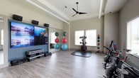 This single-level fitness studio is thoughtfully designed to support a wide range of exercise routines. The room features light-toned hardwood-style flooring that extends throughout, providing a clean and durable surface suited for both cardio and strength training activities. Elevated ceilings with a tray detail add a sense of spaciousness, while a ceiling fan promotes air circulation during intense workouts.

A prominent wall-mounted display system with multiple screens serves as a focal point on one side of the room, accompanied by mounted speakers for an immersive audio-visual experience. A large mirror installed beside the screens enhances the sense of depth and allows for form-checking during exercise. Step platforms are neatly stacked nearby for use in aerobic routines.

Natural light enters through multiple windows fitted with horizontal blinds, brightening the space without harsh glare. The room is well-stocked with exercise equipment, including stability balls arranged on a dedicated rack, a medicine ball storage tower, a dumbbell rack, and a stationary cycling bike. A protective floor mat defines a dedicated training zone in the center of the room, completing this versatile and well-appointed fitness space.