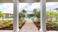 Located at 14050 Integra Dr in Jacksonville, FL, this single-level outdoor amenity space is designed to deliver a resort-like experience within a residential community. The centerpiece of the space is a generously sized swimming pool, framed by towering palm trees and surrounded by lounge chairs, creating a relaxing retreat ideal for the Florida climate.

A herringbone-patterned brick pathway leads visitors from a covered pavilion directly toward the pool area, establishing a clear and inviting sense of arrival. The pavilion itself features crisp white classical columns supporting a covered roof structure, offering shaded outdoor seating with blue Adirondack chairs positioned along the exterior facade. Large windows on the clubhouse building allow natural light to flood the interior while maintaining visual connectivity to the pool and grounds.

The landscaping throughout the space is meticulously maintained, with tropical shrubs, ornamental plants, and mulched garden beds flanking the walkways and structures. A pergola structure is also visible in the background, providing an additional shaded gathering area. The overall ambiance is bright, open, and welcoming, combining classic architectural detailing with vibrant subtropical greenery to create a polished and functional outdoor community hub.