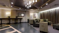 This single-floor space presents a polished and upscale interior designed to impress and provide comfort to its occupants. The room is anchored by a statement chandelier with white globe shades suspended from a recessed, dark-toned ceiling that adds depth and architectural interest overhead. Recessed lighting complements the chandelier, ensuring the space is well-illuminated throughout.

The seating area is furnished with plush, olive-toned upholstered armchairs and loveseats arranged to encourage both relaxed individual seating and conversation. A small counter or bar area along one wall features a sleek surface and a modern bar stool, suggesting a service or reception function. A large framed mirror above this counter adds a sense of openness to the space.

The walls are finished in a textured, neutral tone and adorned with curated artwork, including a notably framed portrait painting that serves as a focal point. Soft wall sconces contribute to the warm, ambient lighting. Flowing floor-length drapery in a muted tone lines one wall, adding an element of elegance and softness. The flooring is a dark, polished surface accented with lighter inlay detailing, grounding the room with a sense of sophistication. Overall, the space conveys a refined, professional atmosphere.