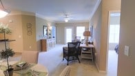 This single-floor space exudes a warm and inviting ambiance through its thoughtfully designed interior. The living area serves as the heart of the home, showcasing elegant crown molding that frames the room and adds a refined architectural touch. Soft, neutral beige tones on the walls complement the plush wall-to-wall carpeting, creating a cohesive and comfortable atmosphere throughout.

Natural light flows generously into the living space through multiple windows and a glass-paneled door, brightening the room and enhancing its open, airy feel. A ceiling fan with a built-in light fixture is centrally positioned, providing both functionality and a classic design element.

The living area is furnished with a mix of wicker and upholstered seating, including a distinctive dark wicker armchair that adds texture and visual interest. A rustic wooden console table along one wall is paired with a warm-toned table lamp, contributing to the cozy, layered lighting scheme. A charming buffet-style credenza with a decorative mirror above it anchors another wall, adding both storage and style.

The dining area, visible in the foreground, features a light-toned table set with place settings, seamlessly connecting the living and dining spaces. Overall, the space offers a harmonious blend of classic detailing and relaxed, livable design.