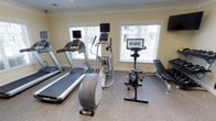 This fitness room offers a clean, functional space designed to support a well-rounded workout routine. The room is outfitted with a selection of cardio equipment, including two treadmills with integrated display screens, an elliptical cross-trainer, and a stationary cycling bike featuring its own dedicated monitor — ideal for guided ride sessions. A weight bench rounds out the strength training options, providing versatility for users of varying fitness levels.

The space is finished with warm yellow walls accented by crisp white crown molding, lending it a polished and inviting atmosphere. Durable, speckled flooring provides a practical surface suited for high-traffic fitness use. Three large windows dressed with white horizontal blinds flood the room with abundant natural light, creating an energizing environment that keeps the space feeling open and bright.

The layout is thoughtfully arranged to maximize usable floor space while ensuring comfortable clearance around each piece of equipment. Whether used for cardio endurance, cycling classes, or strength conditioning, this fitness room delivers a comprehensive and well-maintained environment for an effective workout.