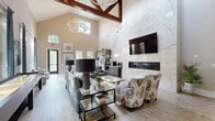Located at 1021 Glenn Valley Lane in Indian Trail, NC, this open-concept kitchen and living room makes a striking impression with its soaring vaulted ceilings accented by bold, exposed dark wood trusses — a defining architectural feature that adds warmth and character to the space. Light wood-look flooring runs throughout, complementing the neutral, warm-toned walls and creating a cohesive, airy atmosphere.

The living area is generously proportioned, bathed in natural light streaming through multiple large windows dressed with elegant teal drapery. A marble-surround linear fireplace serves as a dramatic focal point, framed by floor-to-ceiling porcelain tile. A rustic barn door adds a charming contrast to the otherwise contemporary design. The space comfortably accommodates multiple seating arrangements, ideal for both relaxing and entertaining.

The kitchen is sleek and modern, featuring dark wood cabinetry, a marble-style tile backsplash, and stainless steel appliances. A substantial live-edge communal table anchors the dining area, surrounded by contemporary ghost chairs with gold accents, perfectly balancing rustic and modern aesthetics. Tall windows in the kitchen area flood the space with natural light and offer pleasant outdoor views. A gold-accented bistro table and bar stools provide an additional casual seating option, rounding out this well-appointed, versatile space.