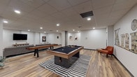 This dynamic single-floor amenity space is designed for recreation and social gathering, offering a well-appointed layout that seamlessly blends game areas with comfortable lounge zones. The room is anchored by a standout pool table with a navy felt surface, positioned atop a bold black-and-white striped area rug, creating a striking visual focal point. Alongside it, a full-length shuffleboard table provides additional gaming options, while a foosball table rounds out the recreational offerings in the space.

A signature "Knick" wall mural serves as a defining decorative element, lending the space a distinctive urban character. Exposed concrete columns add an industrial touch that contrasts warmly with the clean white walls adorned with framed contemporary artwork. The wood-look plank flooring runs throughout, contributing warmth and cohesion across the entire area.

The lounge section features a generously sized gray sectional sofa accented with colorful throw pillows, paired with a woven coffee table set atop a vibrant multi-hued area rug. A large wall-mounted flat-screen TV and a media console complete this relaxation zone. Recessed lighting throughout the drop ceiling ensures even, comfortable illumination across all areas, making this space equally inviting for casual gatherings and competitive play alike.