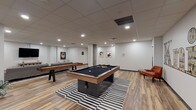 This dynamic single-floor amenity space is designed for recreation and social gathering, offering a well-appointed layout that seamlessly blends game areas with comfortable lounge zones. The room is anchored by a standout pool table with a navy felt surface, positioned atop a bold black-and-white striped area rug, creating a striking visual focal point. Alongside it, a full-length shuffleboard table provides additional gaming options, while a foosball table rounds out the recreational offerings in the space.

A signature "Knick" wall mural serves as a defining decorative element, lending the space a distinctive urban character. Exposed concrete columns add an industrial touch that contrasts warmly with the clean white walls adorned with framed contemporary artwork. The wood-look plank flooring runs throughout, contributing warmth and cohesion across the entire area.

The lounge section features a generously sized gray sectional sofa accented with colorful throw pillows, paired with a woven coffee table set atop a vibrant multi-hued area rug. A large wall-mounted flat-screen TV and a media console complete this relaxation zone. Recessed lighting throughout the drop ceiling ensures even, comfortable illumination across all areas, making this space equally inviting for casual gatherings and competitive play alike.