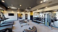 This comprehensive fitness center is thoughtfully designed to accommodate a wide range of workout styles and fitness goals. The space is organized into distinct functional zones, each with its own character and purpose. The main strength training area features a full rack of dumbbells, weight benches, a cable machine with resistance attachments, and a heavy-duty tire for functional training, all set against floor-to-ceiling mirrors that amplify the sense of space. Recessed lighting and ceiling fans on dark-painted ceilings give the area a modern, energetic atmosphere accented by bold yellow walls. A dedicated cardio room houses a row of treadmills positioned in front of large windows that frame open outdoor views, making workouts feel expansive and connected to the outdoors. A separate cycling studio with spin bikes and mirrored walls provides an immersive environment for indoor cycling sessions. A versatile open studio area features a glass roll-up garage door that can open to the outside, a mounted flat-screen TV, and ample floor space for group classes or functional training. This area is stocked with stability balls, medicine balls, kettlebells, resistance bands, and yoga mats. Built-in cubbies near the entrance offer convenient storage for personal belongings, completing this well-rounded facility.