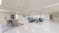 Located at 1080 Vineyard Loop in Iowa, Louisiana, this single-floor space offers a generously proportioned, open-concept layout designed for comfort and versatility. The interior is defined by its bright, neutral palette — crisp white walls and polished light-toned flooring that reflect both natural and artificial light, creating an airy, welcoming atmosphere throughout.

The main gathering area is anchored by a plush blue sofa set arranged around a rustic farmhouse-style coffee table, all set atop a decorative area rug. Multiple windows dressed with sheer linen curtains line the walls, allowing abundant natural light to fill the space. Overhead, flush-mounted linear lighting fixtures provide consistent illumination across the open floor plan.

A dedicated dining area features a wood-toned table with seating for several guests, accompanied by a decorative hutch, contributing a warm, lived-in charm. Adjacent to the main living zone, a compact kitchen area includes cabinetry and appliances, offering functional convenience. A media area with a wall-mounted television and a coordinating media console provides an additional entertainment focal point.

The layout also includes interior doors leading to private restrooms and utility areas, as seen in the floorplan. Built-in storage cabinetry along one wall adds practical functionality. The overall design balances open communal space with purposeful zones, making this a well-appointed and adaptable single-level facility.