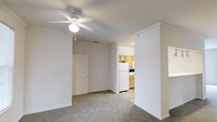 This single-floor residential space offers a clean, functional layout designed for comfortable everyday living. The main living area features neutral-toned carpeting underfoot and smooth white walls that create a bright, open atmosphere. A ceiling fan with an integrated light fixture is centrally mounted, providing both air circulation and ambient lighting to the room. Crown molding along the ceiling adds a subtle architectural detail that elevates the overall finish of the space.

An interior door off the living area provides access to an additional room, contributing to the practical flow of the floor plan. The living space connects openly to the kitchen, which is finished with light wood cabinetry offering ample storage. White appliances, including a refrigerator and range, are neatly integrated into the kitchen layout, which transitions to tile flooring — a durable and easy-to-maintain surface well-suited for a cooking and dining area.

The overall color palette throughout the space is light and neutral, with white walls and ceilings that help maximize the sense of openness. The layout is straightforward and well-organized, making efficient use of the available space. Clean lines, practical finishes, and a cohesive design make this a welcoming and move-in-ready residential space.