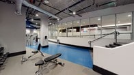 This fitness facility is designed with a functional, industrial-inspired aesthetic that prioritizes usability and open space. The single-floor layout is defined by exposed concrete ceilings with visible ductwork and overhead piping, lending the space an urban, warehouse-style character. Recessed and track lighting throughout ensures consistent illumination across all workout zones.

The main floor features a vibrant blue rubber flooring that defines the primary exercise area, providing cushioning and durability underfoot. This zone is equipped with free weights, adjustable benches, and strength training machines, accommodating a range of workout routines. A structural column anchors the center of the space without disrupting the overall flow of the layout.

A standout feature is the glass-partitioned studio space along one wall, framed by floor-to-ceiling aluminum-framed windows. This enclosed room offers a dedicated area for group classes or focused training, while maintaining visual connectivity with the main floor. Mirrored walls in the strength training area enhance the sense of depth and allow users to monitor their form. The overall ambiance is clean, modern, and purposefully designed for high-performance use.