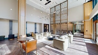 This expansive, single-floor lobby lounge offers a striking first impression through its architectural grandeur and carefully curated design. Soaring double-height ceilings define the main living area, amplifying the sense of openness and volume throughout the space. Floor-to-ceiling glass curtain walls and oversized windows flood the interior with abundant natural light, framing views of an outdoor terrace and the surrounding urban landscape beyond.

The living area is anchored by multiple thoughtfully arranged seating vignettes, featuring plush neutral sofas, leather accent chairs, and round coffee tables set atop layered area rugs over herringbone-patterned hardwood flooring. A built-in wood-paneled feature wall with an integrated fireplace serves as a refined focal point in one seating zone, flanked by warm wood columns that add natural texture and warmth.

A dramatic suspended chandelier installation of cascading pendant lights draws the eye upward, complemented by a tall brass-framed architectural screen partition that elegantly divides areas while maintaining visual continuity. A bar counter with patterned tile detailing and dark bar seating provides a social gathering spot near glass doors that open directly onto the outdoor terrace. The hallway connects seamlessly to additional rooms, maintaining the space's cohesive, contemporary aesthetic throughout.