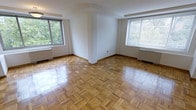This well-appointed single-floor space showcases a thoughtful blend of classic design and comfortable living. The main living area immediately draws attention with its beautifully patterned parquet hardwood flooring, laid in a distinctive herringbone-style arrangement that adds warmth and timeless character to the room. The flooring is finished to a polished sheen, reflecting the natural light that pours in generously through multiple large windows.

The windows are a standout feature, positioned on two walls and framing lush, verdant views of mature trees outside. This abundance of natural light creates an airy, open ambiance throughout the space. Each window is also equipped with a radiator beneath it, indicating a classic heating system that contributes to the building's traditional architectural character.

The walls are finished in clean, crisp white, providing a neutral backdrop that enhances the sense of brightness and space. A structural column or partial wall element is visible near the center of the room, adding an architectural detail that subtly defines areas within the open layout. The overall ambiance is one of understated elegance — a clean, light-filled space with enduring design elements that offer a solid foundation for a variety of interior styles and uses.