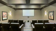 This single-level screening room is designed to deliver a dedicated home theater experience with a polished, purposeful layout. The room is centered around a large retractable projection screen mounted on a neutral-toned wall, flanked by recessed lighting that provides subtle, ambient illumination without interfering with viewing. Dark leather armchairs with built-in cup holders are arranged in two rows, offering clear sightlines to the screen from every seat. A subwoofer positioned at the base of the screen wall anchors the audio setup, suggesting a full surround sound configuration. The ceiling features a structured soffit detail with integrated lighting, adding architectural depth while helping to control acoustics. Striped carpeting runs along the central aisle, providing a clean visual lane between the seating rows. The overall color palette — warm beiges, charcoals, and deep browns — creates a focused, distraction-free environment suited for film screenings, presentations, or private viewings. The space balances function and comfort effectively.