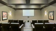 This single-level screening room is designed to deliver a dedicated home theater experience with a polished, purposeful layout. The room is centered around a large retractable projection screen mounted on a neutral-toned wall, flanked by recessed lighting that provides subtle, ambient illumination without interfering with viewing. Dark leather armchairs with built-in cup holders are arranged in two rows, offering clear sightlines to the screen from every seat. A subwoofer positioned at the base of the screen wall anchors the audio setup, suggesting a full surround sound configuration. The ceiling features a structured soffit detail with integrated lighting, adding architectural depth while helping to control acoustics. Striped carpeting runs along the central aisle, providing a clean visual lane between the seating rows. The overall color palette — warm beiges, charcoals, and deep browns — creates a focused, distraction-free environment suited for film screenings, presentations, or private viewings. The space balances function and comfort effectively.