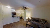 This single-floor space offers a warm and functional open-concept layout that seamlessly connects the living area, dining space, and kitchen. The carpeted living room is anchored by a comfortable sofa and a classic coffee table, creating an inviting area for relaxation. A ceiling fan overhead provides both style and practical air circulation throughout the space. A structural column adds architectural character while subtly defining the transition between the living and dining areas. The kitchen is well-equipped with dark cabinetry, a built-in microwave, and a raised breakfast bar that doubles as a casual dining counter. A pendant light hangs above the bar, adding a touch of warmth to the kitchen area. Adjacent to the kitchen, a compact dining nook with a table and chairs is illuminated by a hanging light fixture, making it ideal for everyday meals. The overall ambiance of the space is cozy and practical, with warm lighting from a table lamp in the living area contributing to a welcoming atmosphere. The layout makes efficient use of the available footprint, ensuring a smooth and natural flow between all areas of the home.