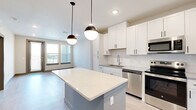 This well-appointed 1-bedroom, 1-bathroom unit offers a thoughtfully designed open-concept layout that maximizes functionality and flow. The combined kitchen and living area serves as the heart of the unit, where a central island with a striking quartz countertop takes center stage, illuminated by two stylish pendant lights. The kitchen is outfitted with white shaker-style cabinetry, subway tile backsplash, and a full suite of stainless steel appliances, including a French door refrigerator, electric range, built-in microwave, and dishwasher. Light wood-style flooring extends seamlessly throughout the entire unit, lending a clean, contemporary feel. The living area benefits from multiple windows that invite natural light and provide outdoor views. The bedroom is a comfortable, well-lit space featuring a ceiling fan and a large window overlooking the surroundings. A dedicated walk-in closet with dual hanging rods and built-in shelving provides generous storage. The bathroom is finished with a wide vanity featuring quartz countertops, white cabinetry with matte black hardware, a large mirror, and a subway tile shower. The unit's neutral color palette and modern finishes create a cohesive and inviting atmosphere throughout.