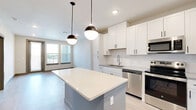 This well-appointed 1-bedroom, 1-bathroom unit offers a thoughtfully designed open-concept layout that maximizes functionality and flow. The combined kitchen and living area serves as the heart of the unit, where a central island with a striking quartz countertop takes center stage, illuminated by two stylish pendant lights. The kitchen is outfitted with white shaker-style cabinetry, subway tile backsplash, and a full suite of stainless steel appliances, including a French door refrigerator, electric range, built-in microwave, and dishwasher. Light wood-style flooring extends seamlessly throughout the entire unit, lending a clean, contemporary feel. The living area benefits from multiple windows that invite natural light and provide outdoor views. The bedroom is a comfortable, well-lit space featuring a ceiling fan and a large window overlooking the surroundings. A dedicated walk-in closet with dual hanging rods and built-in shelving provides generous storage. The bathroom is finished with a wide vanity featuring quartz countertops, white cabinetry with matte black hardware, a large mirror, and a subway tile shower. The unit's neutral color palette and modern finishes create a cohesive and inviting atmosphere throughout.