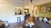 This single-floor residence showcases an open-concept layout that seamlessly connects the kitchen, dining area, and living room into one cohesive living environment. The kitchen features a raised breakfast bar with bar stool seating, warm wood cabinetry, dark countertops, and full-sized appliances including a black refrigerator and stove. A pass-through opening with an arched detail provides a clear sightline into the dining and living areas, enhancing the sense of openness.

The dining area accommodates a round table with dark chairs, positioned beneath a pendant light fixture that adds a focal point overhead. Adjacent to it, the living room is anchored by a plush sofa, a dark wood coffee table, and a decorative accent wall in a rich olive green tone that adds visual depth. An arched niche on the far wall provides an elegant display area for décor.

Throughout the space, rich hardwood-style flooring, crisp crown molding, and a ceiling fan contribute to a polished, comfortable atmosphere. The overall design balances practicality with style in a well-integrated, flowing floor plan.
