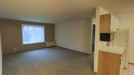 This single-floor space offers a straightforward and functional layout designed for comfortable everyday living. The main living area is generously proportioned, featuring wall-to-wall carpeting that adds warmth underfoot. A large window dressed with vertical blinds allows natural light to fill the room, creating a bright and welcoming atmosphere. Beneath the window sits a through-the-wall HVAC unit, providing both heating and cooling convenience. The walls are finished in a clean, neutral palette — a combination of warm taupe and crisp white — lending the space a calm and versatile aesthetic that can easily accommodate a variety of interior styles. A doorway visible at the rear of the living area leads to an additional room, suggesting a multi-room floor plan. To one side, built-in wooden cabinetry hints at a kitchen or utility area, offering practical storage solutions. The overall ambiance is clean, understated, and move-in ready, with a simple design that prioritizes usability and ease of living. The open feel of the main room provides ample space for furniture arrangement and personalization.