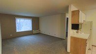This single-floor space offers a straightforward and functional layout designed for comfortable everyday living. The main living area is generously proportioned, featuring wall-to-wall carpeting that adds warmth underfoot. A large window dressed with vertical blinds allows natural light to fill the room, creating a bright and welcoming atmosphere. Beneath the window sits a through-the-wall HVAC unit, providing both heating and cooling convenience. The walls are finished in a clean, neutral palette — a combination of warm taupe and crisp white — lending the space a calm and versatile aesthetic that can easily accommodate a variety of interior styles. A doorway visible at the rear of the living area leads to an additional room, suggesting a multi-room floor plan. To one side, built-in wooden cabinetry hints at a kitchen or utility area, offering practical storage solutions. The overall ambiance is clean, understated, and move-in ready, with a simple design that prioritizes usability and ease of living. The open feel of the main room provides ample space for furniture arrangement and personalization.
