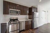 This contemporary single-floor unit showcases a thoughtfully designed open layout with rich, dark hardwood flooring that flows throughout the living areas and bedroom. The kitchen is a standout feature, outfitted with bold, dark wood cabinetry that extends to the ceiling, complemented by a crisp white subway tile backsplash and bright white countertops. A full suite of stainless steel appliances — including a range, over-the-range microwave, dishwasher, and refrigerator — provides a sleek, modern finish. The living area is bathed in natural light and connects seamlessly to the kitchen, offering an airy, open feel. Sliding double doors provide convenient closet access and help define the space efficiently. The bedroom is highlighted by a large floor-to-ceiling window that frames urban views of the surrounding cityscape, flooding the room with natural light and creating an inviting atmosphere. The bathroom features a walk-in shower enclosed by a frameless glass door, lined with clean white subway tile and accented by a small window that introduces additional natural light. Throughout the unit, the clean white walls and modern finishes create a cohesive, contemporary aesthetic that is both functional and visually appealing.