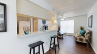 This single-floor residence offers a thoughtfully designed layout within a compact yet efficient footprint. The open-concept living and kitchen area serves as the heart of the home, where a peninsula-style breakfast bar with a light stone countertop creates a natural divide between the kitchen and living space. Two bar stools positioned along the bar make it an ideal spot for casual dining or conversation. A pass-through window connects the kitchen to the living area, enhancing the sense of openness and allowing natural light to flow freely throughout.

The living area features warm hardwood-style flooring that extends throughout the main space, complemented by crisp white walls and a textured ceiling. A ceiling fan is centrally mounted, adding both functionality and a finishing touch to the room. A large window allows abundant natural light to brighten the living area, creating an airy and welcoming ambiance.

The kitchen is visible through the pass-through, offering a clean, bright workspace. Decorative accents throughout the main living area add personality and warmth to the overall design. The space presents a practical and comfortable environment well-suited for everyday living.
