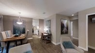 This single-floor residence offers a thoughtfully designed open-concept layout that seamlessly connects the living, dining, and kitchen areas. Rich hardwood-style flooring flows throughout, creating a warm and cohesive aesthetic. The neutral warm-gray walls provide a versatile backdrop, complemented by white baseboards that add a clean, finished look.

The kitchen features crisp white cabinetry, a classic tile backsplash, and stainless steel appliances, providing both functionality and style. A textured ceiling adds subtle character to the space. Recessed lighting and pendant fixtures ensure the interior is well-lit throughout.

The open living and dining area allows for flexible furniture arrangements, as demonstrated by the tasteful staging that includes a cozy seating area with accent pillows and a defined dining space. A small accent desk area serves as a compact workspace or display nook, adorned with decorative pieces and framed artwork. Large-scale wall art in muted, nature-inspired tones adds visual interest and depth to the walls.

A hallway visible from the main living area leads to additional rooms, maintaining a practical and efficient floor plan. Overall, the space conveys a modern, comfortable atmosphere ideal for everyday living.