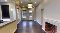 This single-floor space presents a thoughtfully designed interior that balances classic charm with modern touches. The main living area immediately draws attention with its rich, dark hardwood flooring that extends throughout, providing a warm and grounded aesthetic. Neutral warm-gray walls create a versatile backdrop, complemented by crisp white baseboards and trim that add a clean, polished finish.

A standout feature of the living area is the traditional brick fireplace set within a white-painted mantel surround, lending a cozy, timeless character to the room. Overhead, a decorative drum pendant light with a lattice-patterned shade serves as an elegant focal point, while recessed lighting provides additional ambient illumination throughout the space.

Natural light filters into the room through multiple windows fitted with horizontal blinds, offering both brightness and privacy control. A paneled entry door with a transom window further enhances the flow of natural light into the interior.

The layout is open and spacious, with a neatly defined entryway and smooth transitions between areas. Architectural details such as arched or recessed wall niches add subtle visual interest. Overall, the space offers a clean, move-in ready environment with classic design elements that provide lasting appeal and functional comfort.