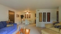 This single-floor residence presents an open and cohesive layout that seamlessly connects the living and dining areas. The living room is furnished with a blue upholstered sofa and a warm wood-toned coffee table accented with decorative candle holders, contributing to a comfortable and inviting atmosphere. Neutral-toned carpeting runs throughout the space, enhancing the sense of continuity between rooms.

The dining area accommodates a glass-topped table surrounded by light-colored upholstered chairs, positioned beneath a ceiling fan with an integrated light fixture. This functional centerpiece provides both ambient lighting and air circulation. Framed artwork adorns the walls throughout, including a grouping of three matching prints in the dining area and a prominent display of tall vertical framed pieces on an accent wall that visually divides the space.

A large indoor plant near the dining area introduces a natural element to the interior. The overall color palette is soft and understated, with cream and beige tones dominating the walls and flooring. The layout is practical and well-organized, with clear sightlines across the main living spaces, making the residence feel open and easy to navigate.