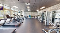 This fitness center at 15350 SW 136th St in Miami, FL is a modern, single-floor gym designed to support a full range of workout routines. The space is defined by its open, expansive layout with a dark rubberized flooring that runs throughout, providing both durability and comfort underfoot. Large floor-to-ceiling windows line multiple walls, flooding the interior with natural light and offering views of the surrounding outdoor grounds, creating an energizing atmosphere for exercise. A suspended drop-tile ceiling with recessed panel lighting ensures consistent illumination throughout the facility. One of the gym's most striking design features is the bold, typographic motivational mural that spans a full accent wall, displaying words such as "Strong," "Believe," "Competitive," "Wellness," and "Achieve," setting an inspirational tone. The cardio section is well-stocked with treadmills, elliptical machines, stationary bikes, and rowing machines. On the opposite side of the floor, a dedicated strength training zone includes a multi-station cable machine, weight benches, a dumbbell rack, and various resistance equipment. A wall-mounted flat-screen television is positioned for visibility during workouts. Storage cubbies and a sanitizing station are also available, adding to the functionality of the space.