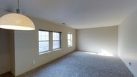 This single-floor residence offers a clean, functional layout designed for comfortable everyday living. The main living area is a standout feature, characterized by its generous proportions, soft wall-to-wall carpeting in a neutral gray tone, and smooth white ceilings that lend the room an open, airy quality. Two double-hung windows along one wall allow natural light to pour in, illuminating the space and creating a warm, welcoming atmosphere. A smaller accent window further enhances the brightness of the room. The walls are finished in a soft, warm beige tone that complements the carpeting and contributes to the overall understated, move-in-ready aesthetic. Subtle architectural details, including a ceiling-mounted light fixture and well-placed electrical outlets, reflect a practical and thoughtfully arranged interior. The open floor plan of the living area provides flexibility for a variety of furniture arrangements and lifestyle needs. The overall ambiance of the space is calm and neutral, offering a versatile blank canvas that can be personalized to suit a range of tastes and preferences.