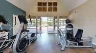 Located at 425 Reflections Cir in San Ramon, CA, this fitness center is housed in a well-appointed single-floor facility defined by its striking vaulted ceiling that rises dramatically overhead, creating an open and airy atmosphere. Large floor-to-ceiling windows and glass double doors flood the space with natural light while offering views of the surrounding greenery and outdoor pool area, giving the room a bright and energizing ambiance.

The interior is finished with durable flooring suited for high-traffic exercise use, and the walls are accented with a deep teal-blue tone that lends a modern, motivating feel. The equipment layout is thoughtfully organized to maximize usable floor space. Cardio machines are prominently featured, including multiple Star Trac treadmills, elliptical cross-trainers, and upright and recumbent stationary bikes. Strength training is also well represented, with at least one dedicated resistance/weight machine visible in the space.

Mounted flat-screen televisions are positioned at multiple points throughout the room, allowing users to stay entertained during their workouts. High clerestory windows near the peak of the vaulted roofline further enhance natural light and ventilation. The space also includes a separate door leading to an additional room, offering potential for further amenities. Overall, the fitness center provides a clean, functional, and well-lit environment suited for a comprehensive workout routine.