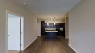 This single-floor apartment offers a thoughtfully designed open-concept layout that seamlessly connects the living area and kitchen. The kitchen is a standout feature, showcasing dark espresso cabinetry that extends to the ceiling for maximum storage, complemented by open shelving for additional display or utility. A central kitchen island provides extra prep space and serves as a natural focal point of the room. The countertops feature a light-toned surface that contrasts elegantly with the dark cabinetry. Black appliances, including a refrigerator, microwave, and range, maintain a cohesive, contemporary aesthetic. Pendant lighting hangs above the island, while a track lighting system illuminates the kitchen workspace, ensuring ample brightness throughout. The flooring throughout the open living and kitchen space consists of rich, dark hardwood-style planks that add warmth and a modern touch. The walls are finished in a neutral warm beige tone, creating a clean and inviting atmosphere. High ceilings enhance the sense of space and airiness throughout the unit. A white paneled door is visible near the entry, adding a classic architectural detail to the otherwise contemporary interior.