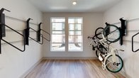 The bike storage room is a thoughtfully designed, single-level space that prioritizes functionality and organization for cyclists. The room features light hardwood-style flooring throughout, creating a clean and easy-to-maintain surface suited to the demands of bicycle storage.

A key feature of the space is the wall-mounted bike rack system, which runs along multiple walls via horizontal metal track rails. Multiple heavy-duty black steel bracket mounts are affixed to these tracks, allowing bikes to be hung vertically and stored efficiently without occupying unnecessary floor space. The system accommodates several bicycles simultaneously and can be adjusted along the track to suit varying storage needs.

Natural light enters the room through a pair of large windows, providing an open, well-lit atmosphere. Recessed ceiling lighting supplements the natural light to ensure visibility throughout the space.

An entryway connects the storage room to an adjacent corridor, which features contrasting dark carpet tile at the transition point and leads toward glazed exterior doors—providing convenient access for bringing bikes in and out. The overall ambiance is clean, practical, and well-suited to active residents seeking secure, organized bicycle storage.