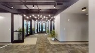This single-floor commercial space presents a refined industrial-chic aesthetic that blends modern design with functional layout. The entrance lobby immediately sets the tone with dramatic globe pendant lighting suspended from an exposed dark ceiling that highlights structural ductwork and mechanical systems — a deliberate design choice that adds urban character. Dark-framed glass doors and sidelights frame the entry points, allowing natural light from the outdoors to filter in and create an open, welcoming atmosphere.

The lobby floor features a combination of patterned carpet tiles and smooth flooring, delineating pathways and gathering areas. Lush potted plants are positioned throughout, adding warmth and a touch of nature to the otherwise sleek interior. Clean white walls provide a neutral backdrop, enhanced by wall-mounted linear light fixtures that offer consistent, even illumination.

A textured wave-panel accent wall adds a tactile focal point near the elevator lobby, where a stainless steel elevator and recessed pendant lighting contribute to a polished, professional feel. Beyond the main lobby, a long corridor extends the full depth of the building, lined with multiple private rooms accessible through individual doors — ideal for offices, studios, or consultation spaces. The consistent use of carpet tiles, neutral tones, and overhead lighting throughout creates a cohesive and professional environment.