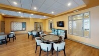 Located in Ramsey, Minnesota, this single-floor game room offers a well-appointed recreational space designed for social gatherings and leisure activities. The room is finished with warm golden-yellow walls and durable hardwood-style flooring throughout, creating an inviting and energetic atmosphere.

The space is divided into distinct activity zones. A dedicated card and game area features multiple round poker tables surrounded by comfortable upholstered chairs, providing ample seating for group play. A wall-mounted flat-screen television and a decorative display cabinet add functional and aesthetic touches to this zone. A Scrabble board display and framed card suit artwork reinforce the gaming theme throughout.

Toward the rear of the room, a full-size billiards table takes center stage, illuminated by a stylish pendant light fixture. Cue racks are mounted nearby for convenient access. Large windows with horizontal blinds line the walls in this section, allowing generous natural light to filter in while offering views of the surrounding exterior.

The drop-panel ceiling features recessed lighting throughout, ensuring consistent, even illumination across the entire space. A casual seating area near the windows provides a quieter spot for conversation. The overall layout is open and well-organized, making this game room a versatile and functional amenity.