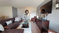 Located at 3526 Langrehr Road in Windsor, MD, this thoughtfully designed single-floor residence offers a cohesive and stylish living experience throughout its well-appointed spaces. The living room serves as the heart of the home, featuring warm neutral carpeting, gray accent walls, and an abundance of natural light flowing in through large vertical-blind-covered windows. Rich brown leather seating, a glass-top coffee table set atop a plush faux-fur rug, and a dark wood media credenza give the space a sophisticated, layered aesthetic. A decorative sunburst mirror adds an artful focal point to the room. The dining area, conveniently connected to the kitchen, features a round wooden dining table surrounded by upholstered chairs, soft carpeting, and a ceiling fan overhead. Wall art and a display cabinet contribute to the room's warm, inviting atmosphere. The kitchen is equipped with dark cabinetry, granite-style countertops, and black appliances including a full-size refrigerator and microwave. The bedrooms are generously proportioned, with one showcasing a bold teal accent wall, a large window with ample natural light, and plush carpeting, while another features a classic four-poster bed and ladder-style shelving, creating a cozy and functional retreat.