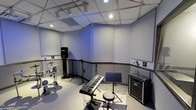 This dedicated music rehearsal studio is purpose-built for musicians seeking a professional, acoustically controlled environment. The room features wall-to-wall acoustic panel treatment across both the walls and drop ceiling, effectively minimizing sound reflection and ensuring a clean, controlled sound experience. The ceiling incorporates a combination of recessed lighting and track lighting, with accent lighting adding a cool blue ambiance to the space.

The studio is equipped to accommodate a full band setup, as evidenced by the drum kit positioned in one corner, a full-size keyboard with stand at the center, a music stand, and a Peavey guitar amplifier. A PA speaker on a stand provides monitoring capability. Multiple power outlets are distributed along the walls, offering convenient access for instrument and equipment connections.

The walls are constructed of painted concrete block, lending the space a solid, sound-isolating quality, while the light grey tones throughout keep the environment bright and focused. The layout is thoughtfully arranged to allow musicians to position themselves comfortably during rehearsals. Overall, this studio offers a functional, well-equipped, and acoustically optimized setting ideal for band practice, songwriting sessions, or individual instrument rehearsal.